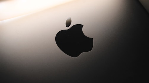 A close-up view of a sleek metallic surface featuring a prominent, glossy black apple-shaped logo with a bite taken out of its right side. The background is softly illuminated, creating a modern and minimalist aesthetic.