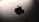 A close-up view of a sleek metallic surface featuring a prominent, glossy black apple-shaped logo with a bite taken out of its right side. The background is softly illuminated, creating a modern and minimalist aesthetic.