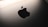 A close-up view of a sleek metallic surface featuring a prominent, glossy black apple-shaped logo with a bite taken out of its right side. The background is softly illuminated, creating a modern and minimalist aesthetic.