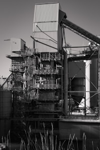 A large industrial structure with multiple interconnected metal pipes and storage units, featuring angular and cylindrical shapes. The image depicts a complex assembly of platforms, ladders, and scaffolding arranged around rectangular and cylindrical components.