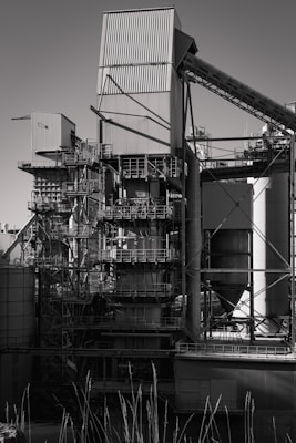 A large industrial structure with multiple interconnected metal pipes and storage units, featuring angular and cylindrical shapes. The image depicts a complex assembly of platforms, ladders, and scaffolding arranged around rectangular and cylindrical components.