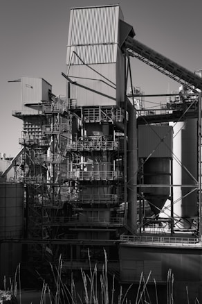 A large industrial structure with multiple interconnected metal pipes and storage units, featuring angular and cylindrical shapes. The image depicts a complex assembly of platforms, ladders, and scaffolding arranged around rectangular and cylindrical components.