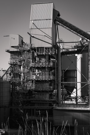 A large industrial structure with multiple interconnected metal pipes and storage units, featuring angular and cylindrical shapes. The image depicts a complex assembly of platforms, ladders, and scaffolding arranged around rectangular and cylindrical components.