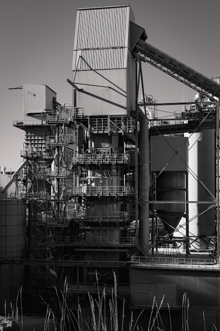 A large industrial structure with multiple interconnected metal pipes and storage units, featuring angular and cylindrical shapes. The image depicts a complex assembly of platforms, ladders, and scaffolding arranged around rectangular and cylindrical components.