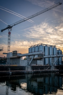 A large construction site features several industrial structures including tall, cylindrical storage tanks and scaffolding. A crane towers over the site, reaching towards the sky. The twilight sky is filled with streaks of clouds illuminated by the setting sun, which casts reflections on the water in the foreground.