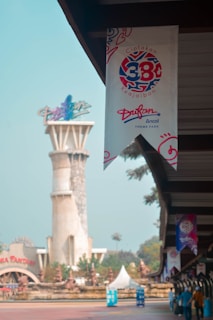 A theme park setting with a prominent banner featuring a logo and text in a foreign language. In the background, there's a tall tower-like structure and various architectural elements, along with greenery and palm trees. The atmosphere appears lively yet calm, with clear skies and daylight.