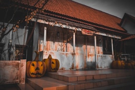 A rustic, old building with a tiled roof and a weathered facade. The porch is adorned with several carved pumpkins, giving it a Halloween theme. Vines and dried leaves hang from the structure, adding to the eerie atmosphere. A statue resembling a ghost or ghoul is positioned to the side, enhancing the spooky vibe.