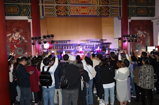 A large crowd gathers indoors facing a performance stage. The stage features traditional musical instruments like drums and bell chimes, and performers dressed in cultural attire. The audience captures the moment on their phones amidst ornate wall decorations and wooden architectural details.