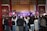 A large crowd gathers indoors facing a performance stage. The stage features traditional musical instruments like drums and bell chimes, and performers dressed in cultural attire. The audience captures the moment on their phones amidst ornate wall decorations and wooden architectural details.