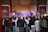 A large crowd gathers indoors facing a performance stage. The stage features traditional musical instruments like drums and bell chimes, and performers dressed in cultural attire. The audience captures the moment on their phones amidst ornate wall decorations and wooden architectural details.