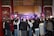 A large crowd gathers indoors facing a performance stage. The stage features traditional musical instruments like drums and bell chimes, and performers dressed in cultural attire. The audience captures the moment on their phones amidst ornate wall decorations and wooden architectural details.