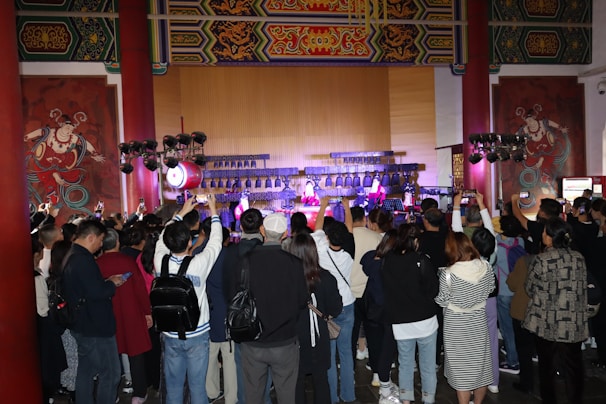 A large crowd gathers indoors facing a performance stage. The stage features traditional musical instruments like drums and bell chimes, and performers dressed in cultural attire. The audience captures the moment on their phones amidst ornate wall decorations and wooden architectural details.