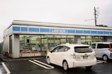 A convenience store with a blue and white color scheme, branded with the name 'LAWSON'. The front windows display various advertisements and signs. Two cars are parked in the lot, including a white Toyota Prius. The sky is overcast, and a utility pole is visible in the background.