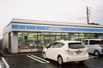 A convenience store with a blue and white color scheme, branded with the name 'LAWSON'. The front windows display various advertisements and signs. Two cars are parked in the lot, including a white Toyota Prius. The sky is overcast, and a utility pole is visible in the background.