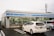 A convenience store with a blue and white color scheme, branded with the name 'LAWSON'. The front windows display various advertisements and signs. Two cars are parked in the lot, including a white Toyota Prius. The sky is overcast, and a utility pole is visible in the background.