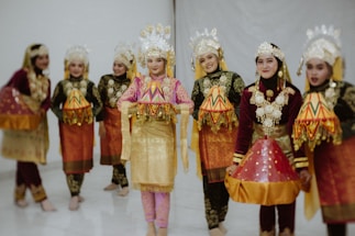 A group of people dressed in traditional costumes, featuring ornate headdresses and embroidered garments. They are wearing vibrant fabrics with gold accents and intricate patterns. Their attire suggests cultural significance, likely related to a traditional dance or ceremony.