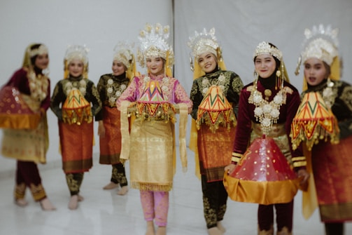 A group of people dressed in traditional costumes, featuring ornate headdresses and embroidered garments. They are wearing vibrant fabrics with gold accents and intricate patterns. Their attire suggests cultural significance, likely related to a traditional dance or ceremony.