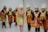 A group of people dressed in traditional costumes, featuring ornate headdresses and embroidered garments. They are wearing vibrant fabrics with gold accents and intricate patterns. Their attire suggests cultural significance, likely related to a traditional dance or ceremony.