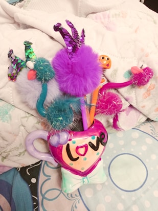 A colorful mug with the word 'love' written on it holds several fuzzy novelty pens with bright pom-poms and sequins. The pens have different colors such as purple, teal, and pink. The background consists of a soft, patterned fabric with pastel shades.