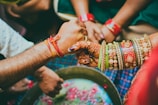 Two people are engaging in a traditional ritual or ceremony. Their hands are adorned with colorful bangles and bracelets, with one person having intricate henna designs on their hand. They appear to be holding or grasping each other's hands over a circular bowl with flower petals.