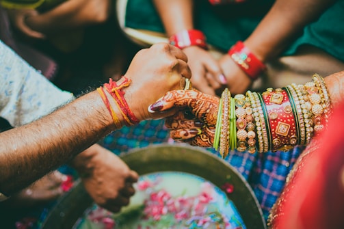 Two people are engaging in a traditional ritual or ceremony. Their hands are adorned with colorful bangles and bracelets, with one person having intricate henna designs on their hand. They appear to be holding or grasping each other's hands over a circular bowl with flower petals.