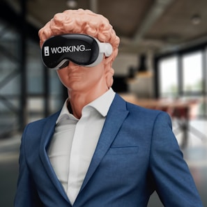 A classical statue-like figure with curly reddish hair is wearing a modern blue suit and a white shirt. The figure is also donning a virtual reality headset displaying the word 'WORKING...'. The setting appears to be a contemporary office or workspace with glass windows in the background.