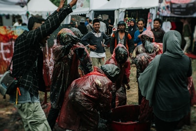 A group of people, some wearing raincoats or protective gear, appear to be engaged in an outdoor event or activity. The scene is set in a crowded festival or market area with tents in the background. The ground is muddy, suggesting recent rain, and one person appears to be raising their arm enthusiastically.