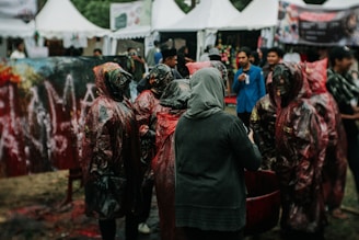 A group of individuals, some wearing protective gear possibly for a paint-related activity, gather outdoors in a festival-like setting. Tents in the background suggest a fair or public event. The scene is busy with people interacting.