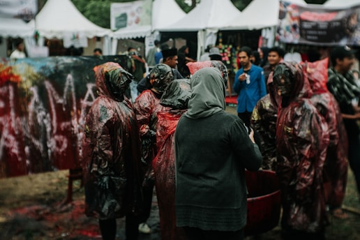 A group of individuals, some wearing protective gear possibly for a paint-related activity, gather outdoors in a festival-like setting. Tents in the background suggest a fair or public event. The scene is busy with people interacting.