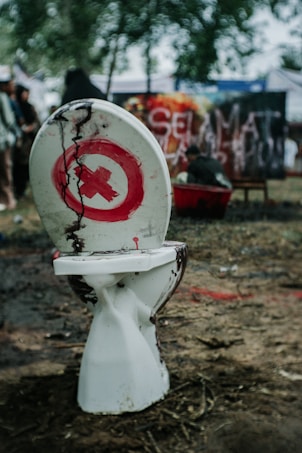 A broken white toilet sits outdoors on earthy ground. The toilet seat and lid are stained with dark, muddy streaks, and there is a red circle with a cross inside painted on the lid. In the background, there are blurry figures and a graffiti wall with partially visible writing. Trees can be seen behind, giving a sense of an outdoor setting.