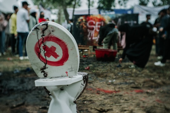 A cracked and damaged toilet with a red cross symbol painted on its lid is placed on a dirt ground. In the background, a group of people gather around what seems to be an art display or installation, some of them bent over examining or interacting with objects.