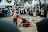 A person wearing a red rain poncho is pulling another person out of a red tub filled with mud. The event is taking place outdoors, surrounded by a large group of people watching. Some onlookers are taking photos. A toilet is placed nearby in the background.