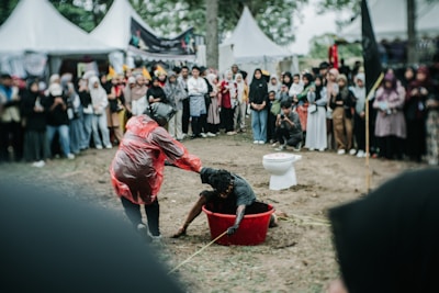 A person wearing a red rain poncho is pulling another person out of a red tub filled with mud. The event is taking place outdoors, surrounded by a large group of people watching. Some onlookers are taking photos. A toilet is placed nearby in the background.