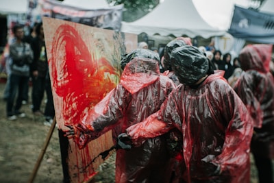 Several people dressed in red plastic ponchos and black hoods appear engaged in a creative activity, seemingly splattering bright red and other colors on a canvas. Surrounding them are others in a crowd, some of whom are observing. The setting appears to be outdoors, perhaps at an art festival or event, indicated by tents and people gathered around.
