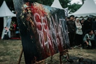 A large abstract painting with bold strokes of black, red, and hints of white sits on an easel outdoors. A group of people stand around it, appearing to be at an art event or festival. There are distinctive white letters smeared across the canvas. In the background, white tent structures can be seen alongside trees.