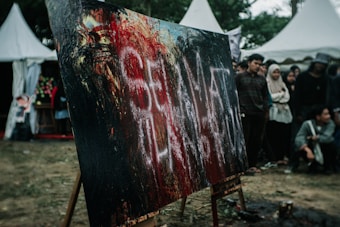A large abstract painting with bold strokes of black, red, and hints of white sits on an easel outdoors. A group of people stand around it, appearing to be at an art event or festival. There are distinctive white letters smeared across the canvas. In the background, white tent structures can be seen alongside trees.