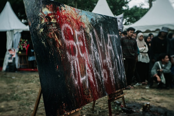 A large abstract painting with bold strokes of black, red, and hints of white sits on an easel outdoors. A group of people stand around it, appearing to be at an art event or festival. There are distinctive white letters smeared across the canvas. In the background, white tent structures can be seen alongside trees.