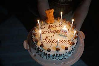 A birthday cake with candles lit, featuring a small teddy bear figurine on top. The cake is decorated with icing and chocolates, and has a message written in a foreign language. There is a large number '6' candle indicating the age.