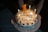 A birthday cake with candles lit, featuring a small teddy bear figurine on top. The cake is decorated with icing and chocolates, and has a message written in a foreign language. There is a large number '6' candle indicating the age.