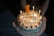 A birthday cake with candles lit, featuring a small teddy bear figurine on top. The cake is decorated with icing and chocolates, and has a message written in a foreign language. There is a large number '6' candle indicating the age.