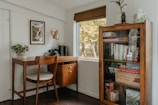 A cozy home office corner features a wooden desk with a vintage rotary phone, a potted plant, and a chair. Above the desk is a framed piece of art and a brass wall lamp. Next to the desk, a tall cabinet with a glass door is filled with books, board games, and decorative items. A window with a wooden blind overlooks a view of trees. The ambiance is warm and inviting, with a mix of modern and rustic elements.