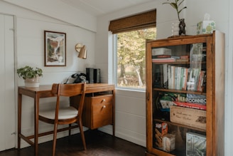 A cozy home office corner features a wooden desk with a vintage rotary phone, a potted plant, and a chair. Above the desk is a framed piece of art and a brass wall lamp. Next to the desk, a tall cabinet with a glass door is filled with books, board games, and decorative items. A window with a wooden blind overlooks a view of trees. The ambiance is warm and inviting, with a mix of modern and rustic elements.