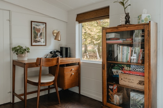 A cozy home office corner features a wooden desk with a vintage rotary phone, a potted plant, and a chair. Above the desk is a framed piece of art and a brass wall lamp. Next to the desk, a tall cabinet with a glass door is filled with books, board games, and decorative items. A window with a wooden blind overlooks a view of trees. The ambiance is warm and inviting, with a mix of modern and rustic elements.