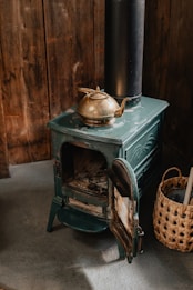 A vintage green cast iron wood stove stands on a concrete floor. A brass kettle sits atop the stove, enhancing its antique appearance. The stove door is open, revealing the interior. To the right, a woven basket contains various items, adding a rustic touch. Wooden walls in the background create a cozy, cabin-like atmosphere.