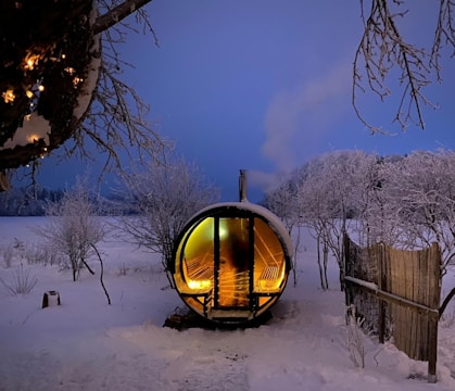 A snow-covered landscape with a cozy, wooden barrel sauna emitting warm light from its interior. Surrounding the sauna are frost-laden trees and sparse fencing, creating a serene, wintery scene. The sky is a soft twilight blue, adding to the tranquil ambiance.