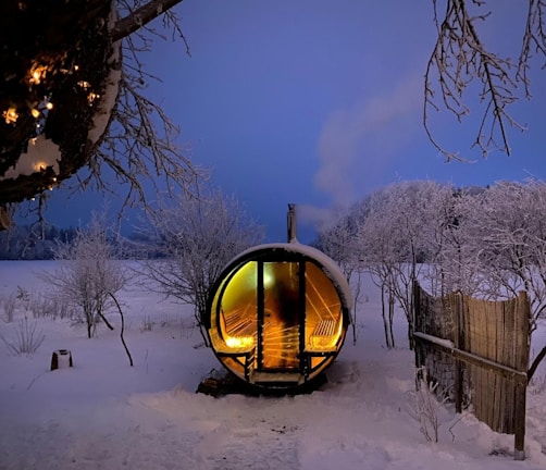 A snow-covered landscape with a cozy, wooden barrel sauna emitting warm light from its interior. Surrounding the sauna are frost-laden trees and sparse fencing, creating a serene, wintery scene. The sky is a soft twilight blue, adding to the tranquil ambiance.