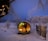 A snow-covered landscape with a cozy, wooden barrel sauna emitting warm light from its interior. Surrounding the sauna are frost-laden trees and sparse fencing, creating a serene, wintery scene. The sky is a soft twilight blue, adding to the tranquil ambiance.