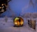 A snow-covered landscape with a cozy, wooden barrel sauna emitting warm light from its interior. Surrounding the sauna are frost-laden trees and sparse fencing, creating a serene, wintery scene. The sky is a soft twilight blue, adding to the tranquil ambiance.
