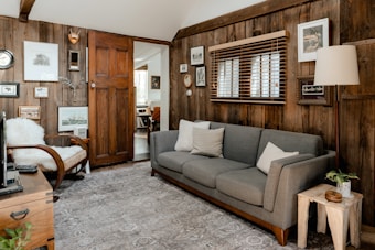 A cozy living room features wooden paneling decorated with various framed artworks. A gray sofa accompanied by white and beige pillows sits along one wall. A wooden chair with a white fluffy blanket is positioned nearby. A small wooden side table with a potted plant and a lamp is next to the sofa, while blinds cover a window above. The floor is covered with a patterned rug, and a glimpse of an adjacent room through an open door reveals more decor.