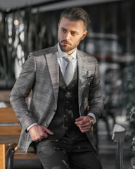 A man dressed in a stylish checkered blazer and vest stands confidently with one hand in his pocket. He is in a modern, possibly indoor setting with blurred background elements such as chairs and tables. The lighting casts shadows and highlights on his face and suit.